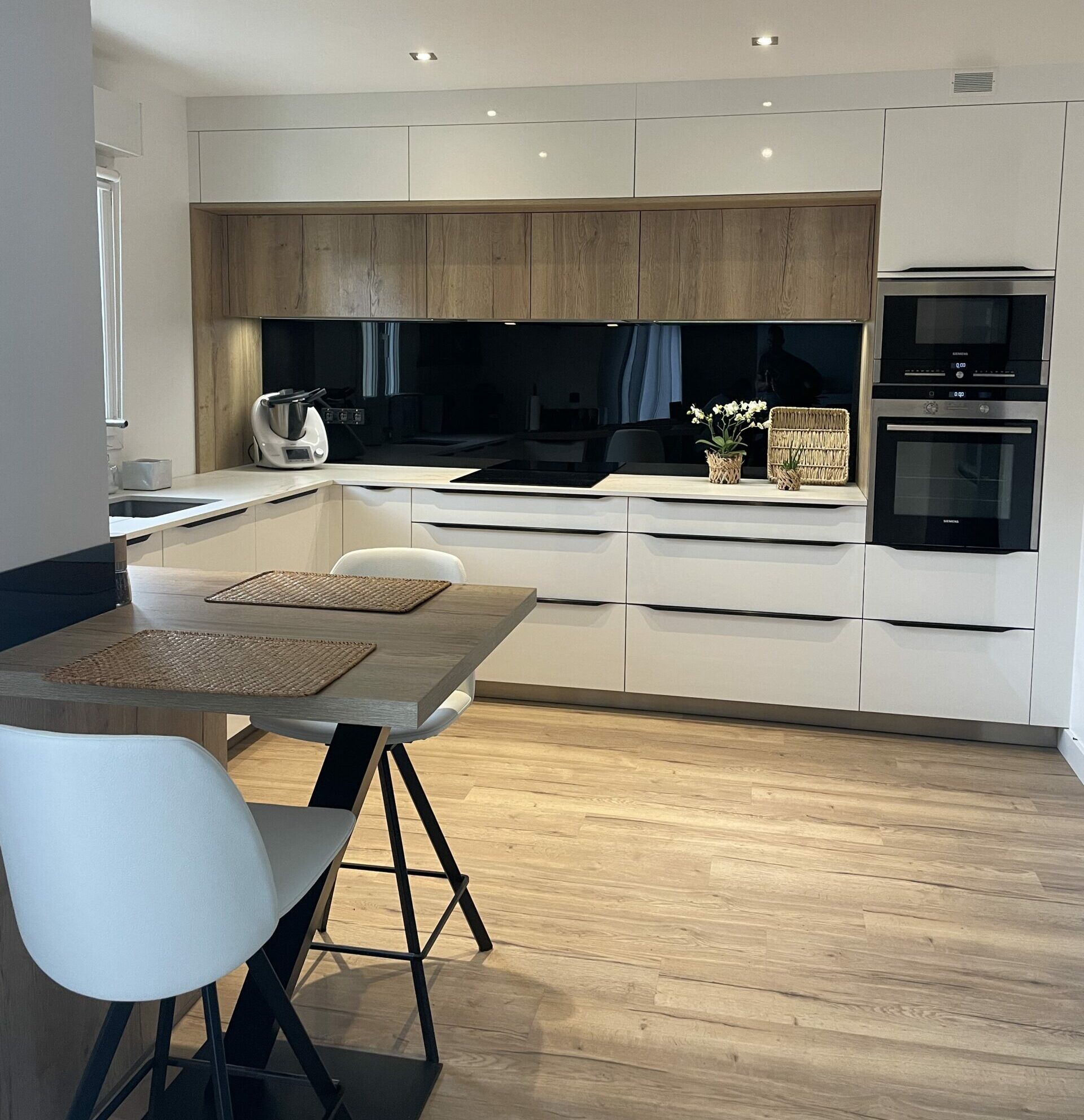 Cuisine contemporaine avec façades blanches mates, éléments bois, crédence noire et électroménager encastré.