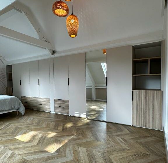 Dressing sur mesure sous combles avec façades blanches, finitions bois, miroir intégré et rangements adaptés aux volumes de la pièce.